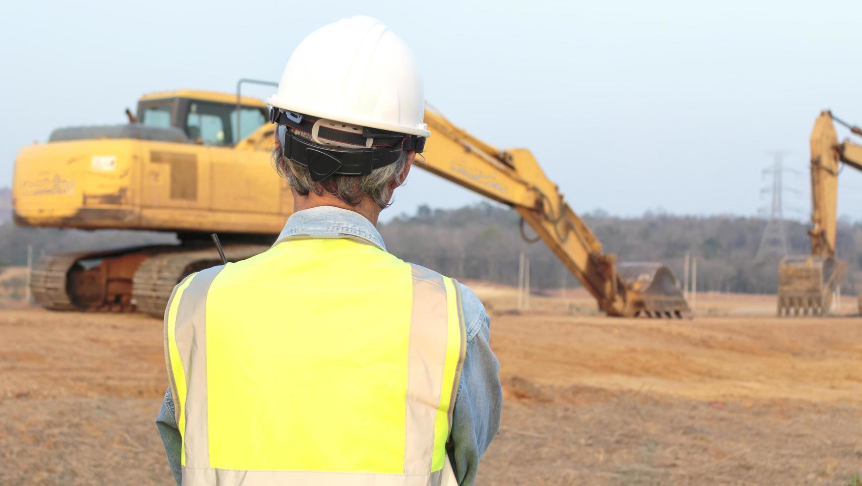 Rear view of a senior Asian male engineer wearing safety shirt and helmet inspecting civil works. The Chief Construction Engineer controls the excavator. photo