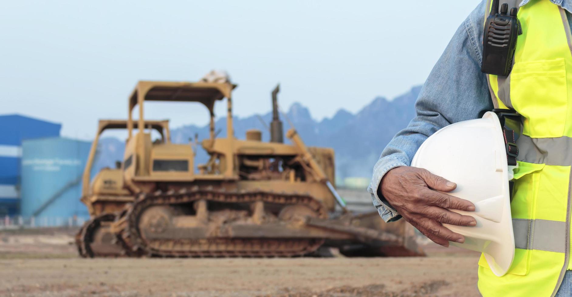 An engineer or worker wearing a safety vest holds a radio transmitter and a white helmet in his hands. for safety working on sunset background and crawler bulldozer photo