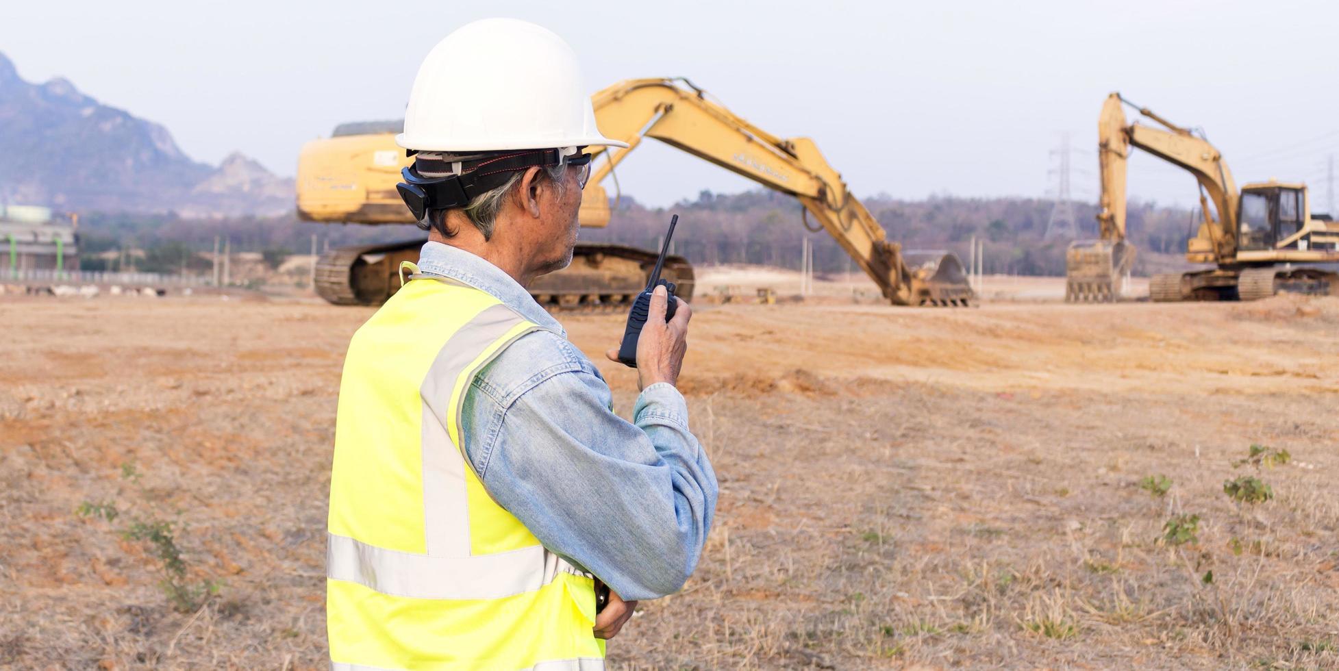 vista lateral, ingeniero senior masculino asiático con chaleco de seguridad y casco inspecciona obras civiles. el ingeniero jefe de construcción usa un walkie-talkie para manejar la excavadora. foto