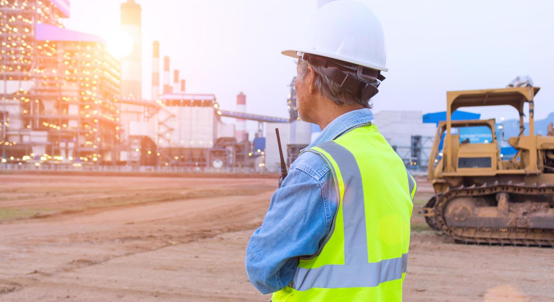 An engineer or worker wearing a safety vest holds a radio transmitter and a white helmet in his hands. for safety working on sunset background and crawler bulldozer photo