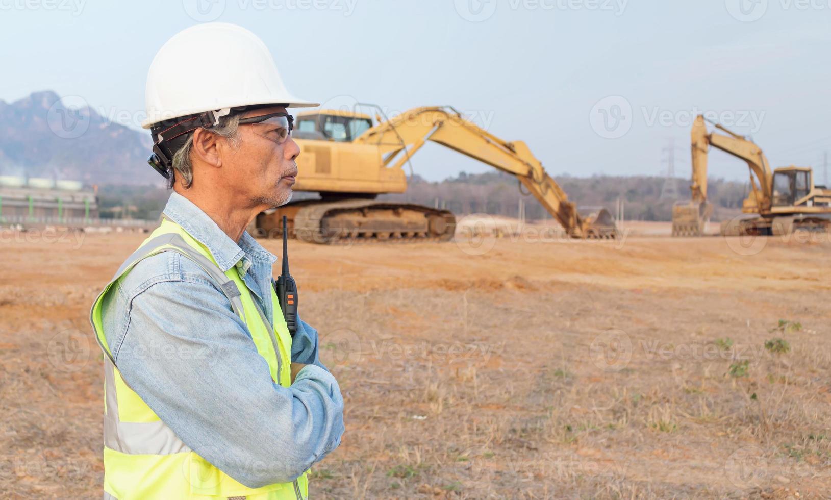 vista lateral ingeniero senior masculino asiático con chaleco y casco inspecciona las obras civiles, el capataz de mediana edad controla la excavadora. foto