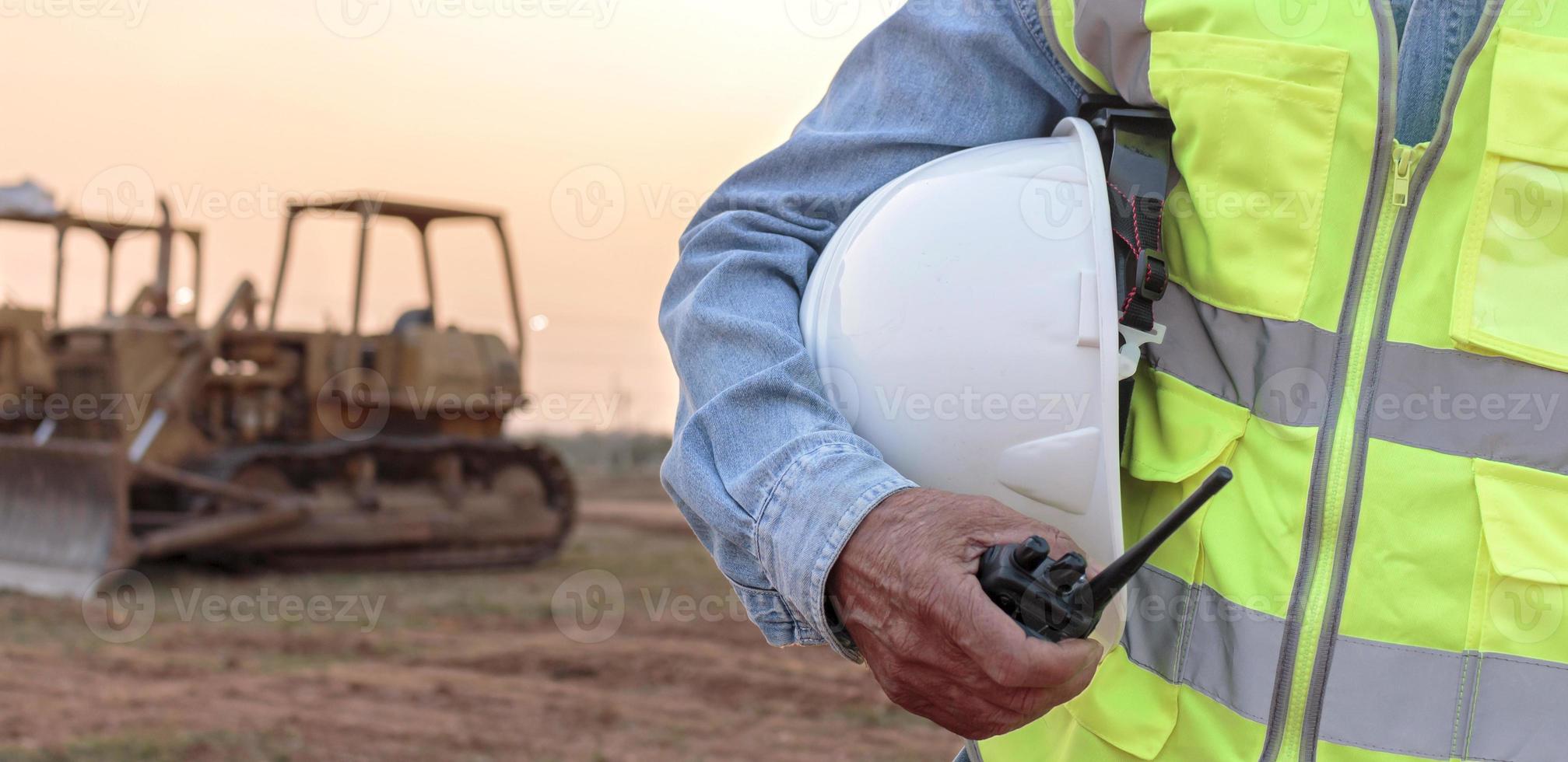 un ingeniero o trabajador que usa un chaleco de seguridad sostiene un transmisor de radio y un casco blanco en sus manos. para la seguridad trabajando en el fondo de la puesta del sol y la excavadora sobre orugas foto