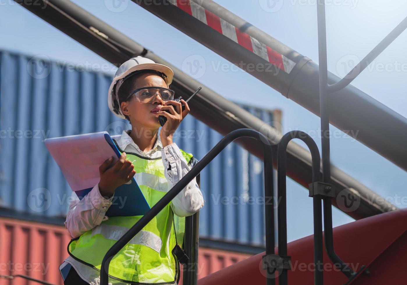 Woman Foreman in hardhat and safety vest holding holding clipboard checklist and talks on two-way radio control loading containers box from cargo photo
