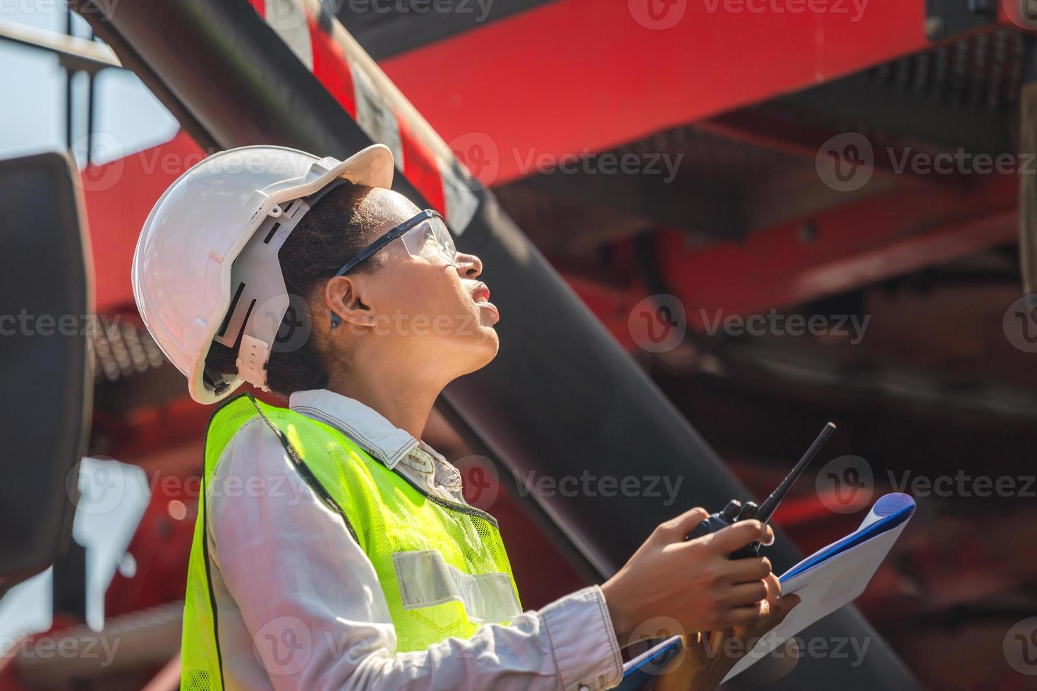 Woman Foreman in hardhat and safety vest holding holding clipboard checklist and talks on two-way radio control loading containers box from cargo photo