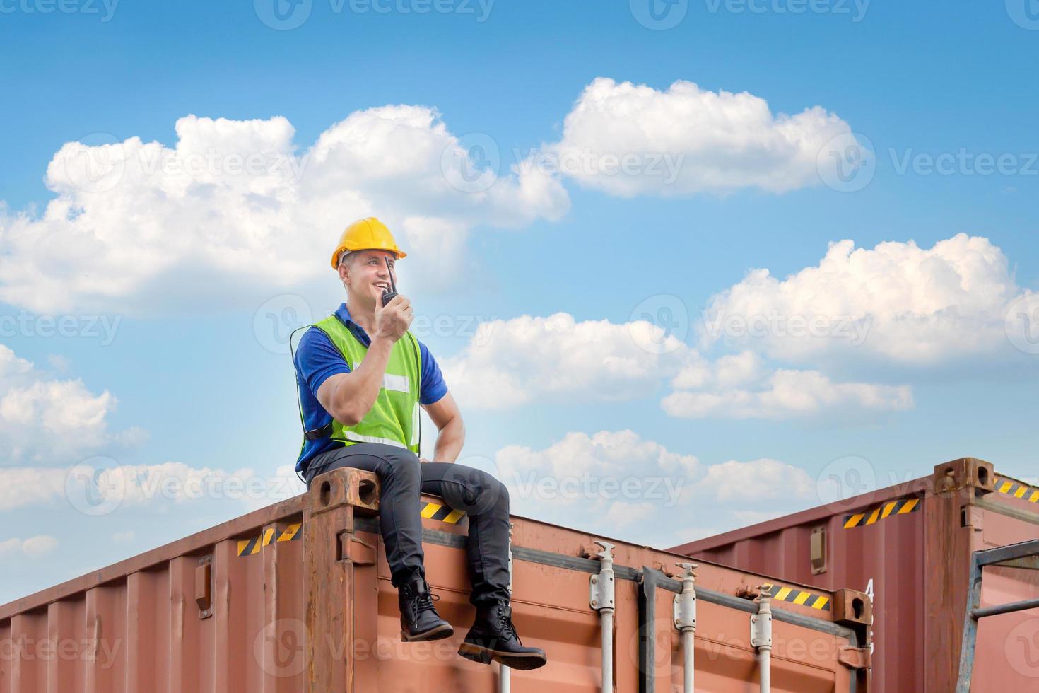 Foreman worker in hardhat and safety vest sitting on container box talks on two-way radio for control loading containers box from cargo photo