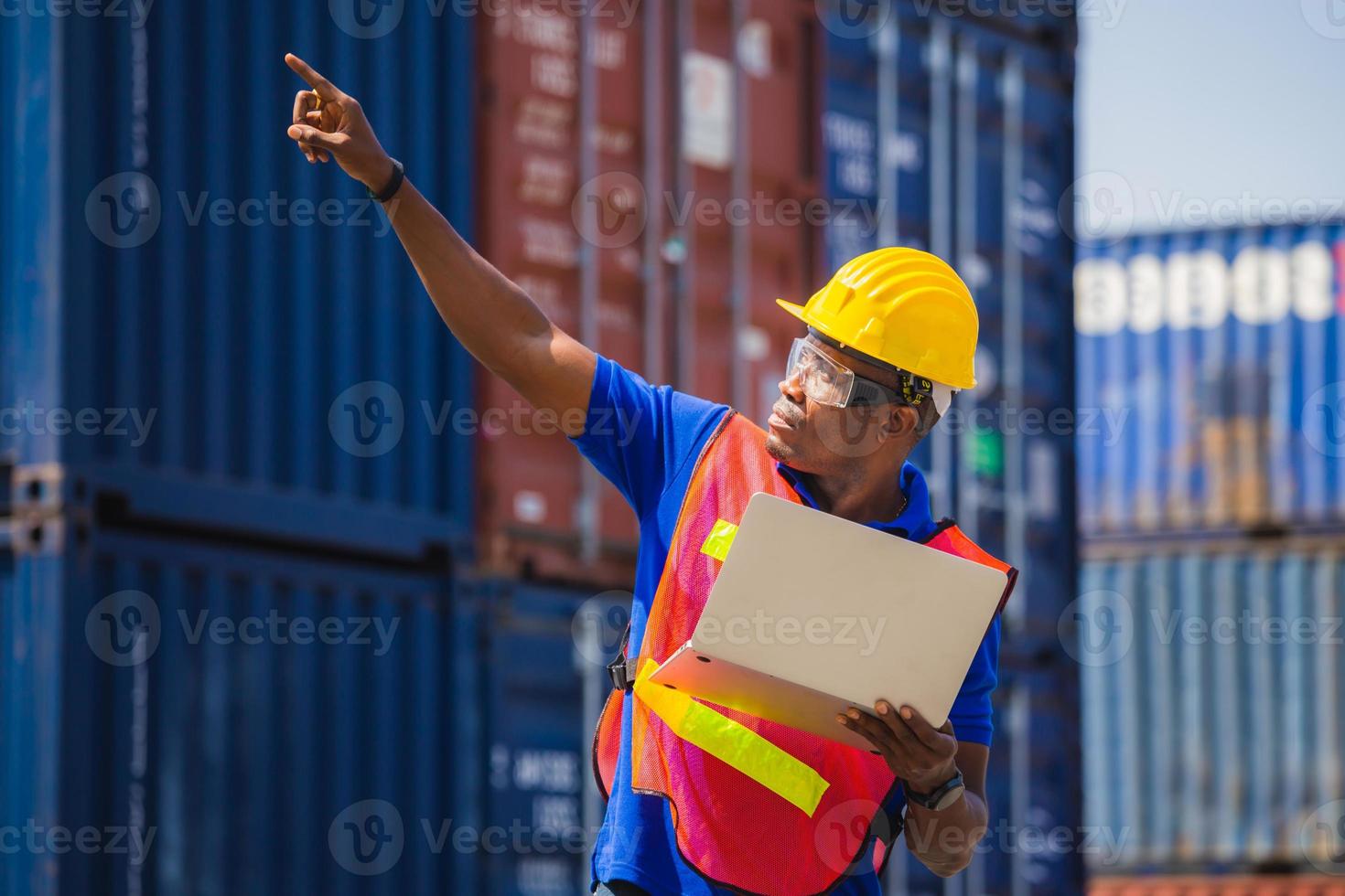 hombre trabajador con casco y chaleco de seguridad que sostiene una computadora portátil y apunta al cielo, el capataz controla la carga de la caja de contenedores de la carga foto