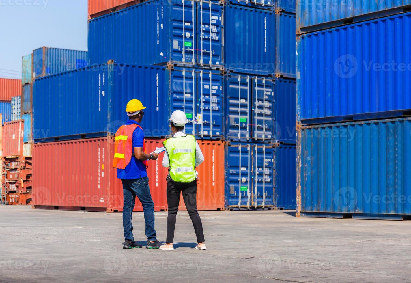 Female foreman with safety vest holding laptop and  worker man in hardhat checking and control loading containers box from cargo photo