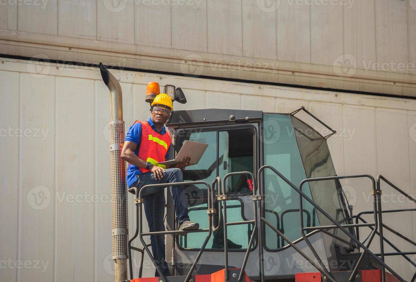 Worker man in hardhat and safety vest holding laptop standing on container stackers control loading containers box from cargo photo