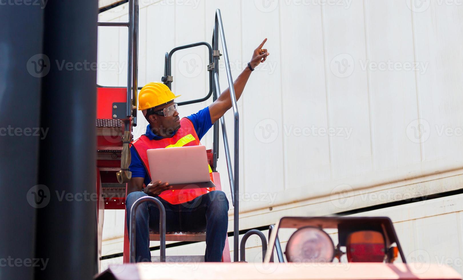 Worker man in hardhat and safety vest holding laptop on container stackers control loading containers box from cargo photo