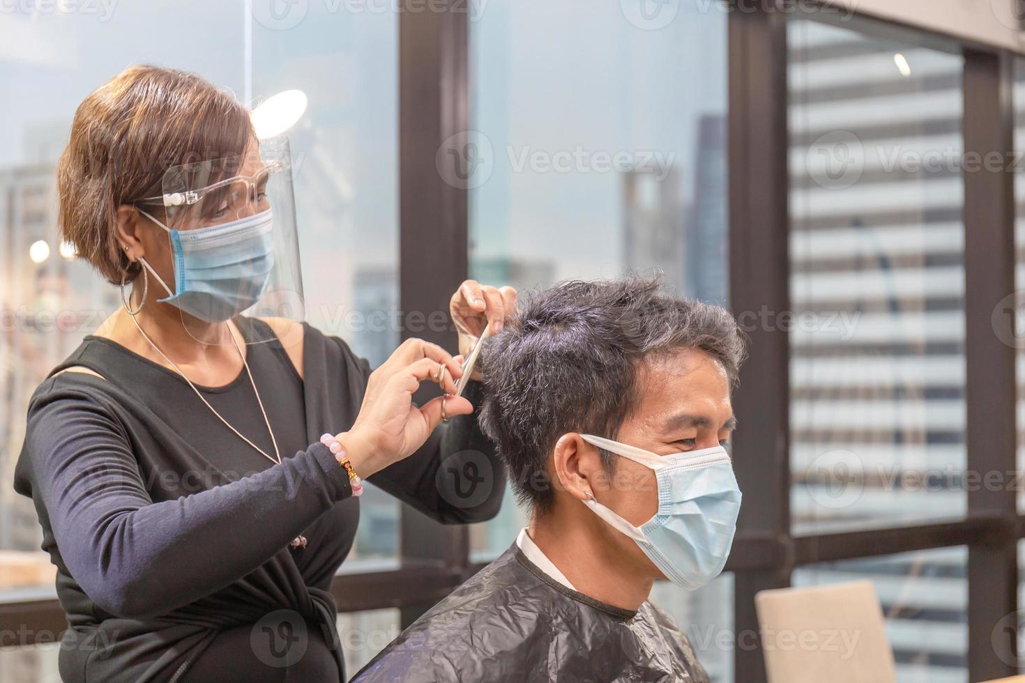 joven que se corta el pelo con peluquero, peluquero con tijeras y peine, peluquero y cliente con máscara protectora debido a la pandemia del coronavirus, nuevos conceptos normales foto