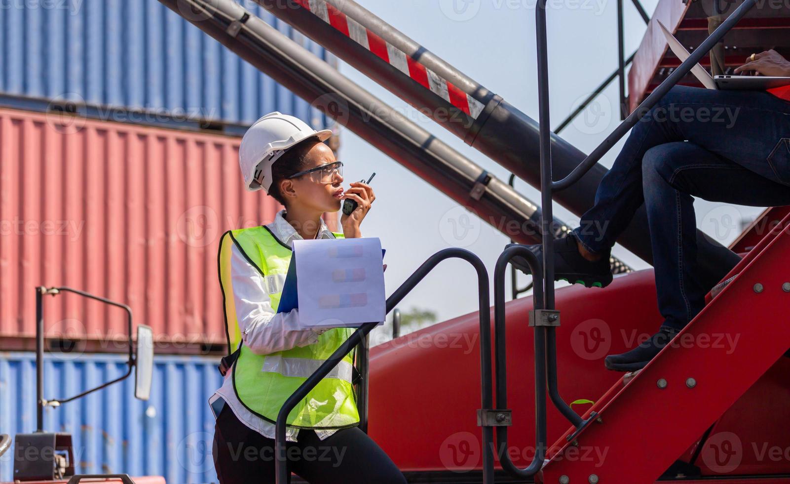 Woman Foreman in hard hat and safety vest holding clipboard checklist and talks on two-way radio, Engineer team control loading containers box from cargo photo