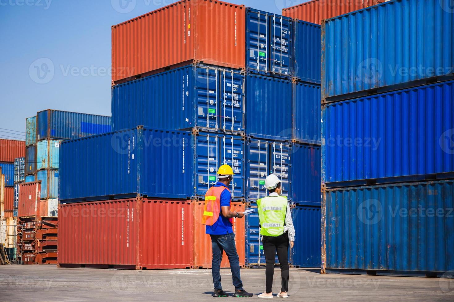 Female foreman with safety vest holding laptop and  worker man in hardhat checking and control loading containers box from cargo photo
