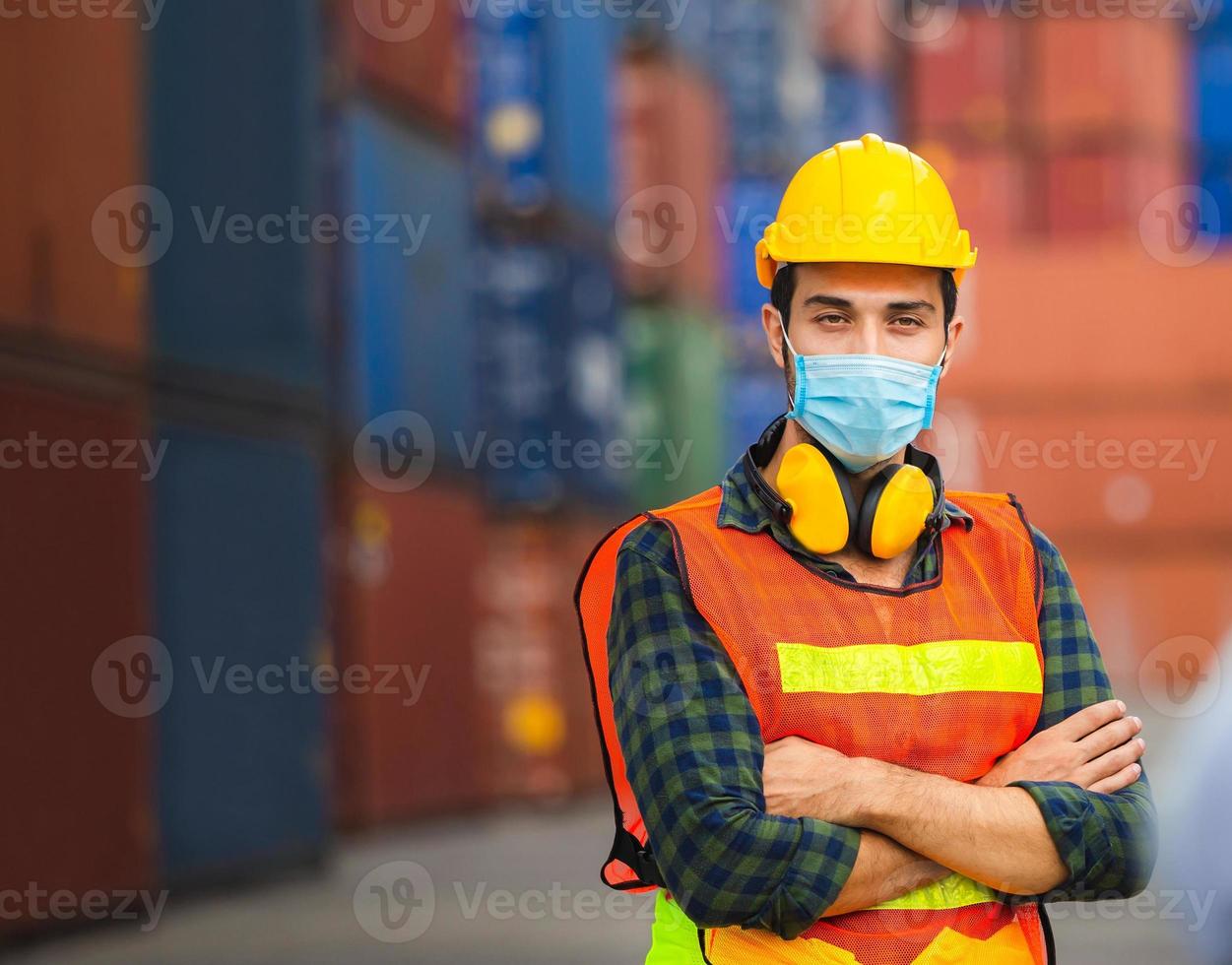 ingeniero alegre con sombrero duro con mascarilla de protección contra el coronavirus con los brazos cruzados en el contenedor de carga foto