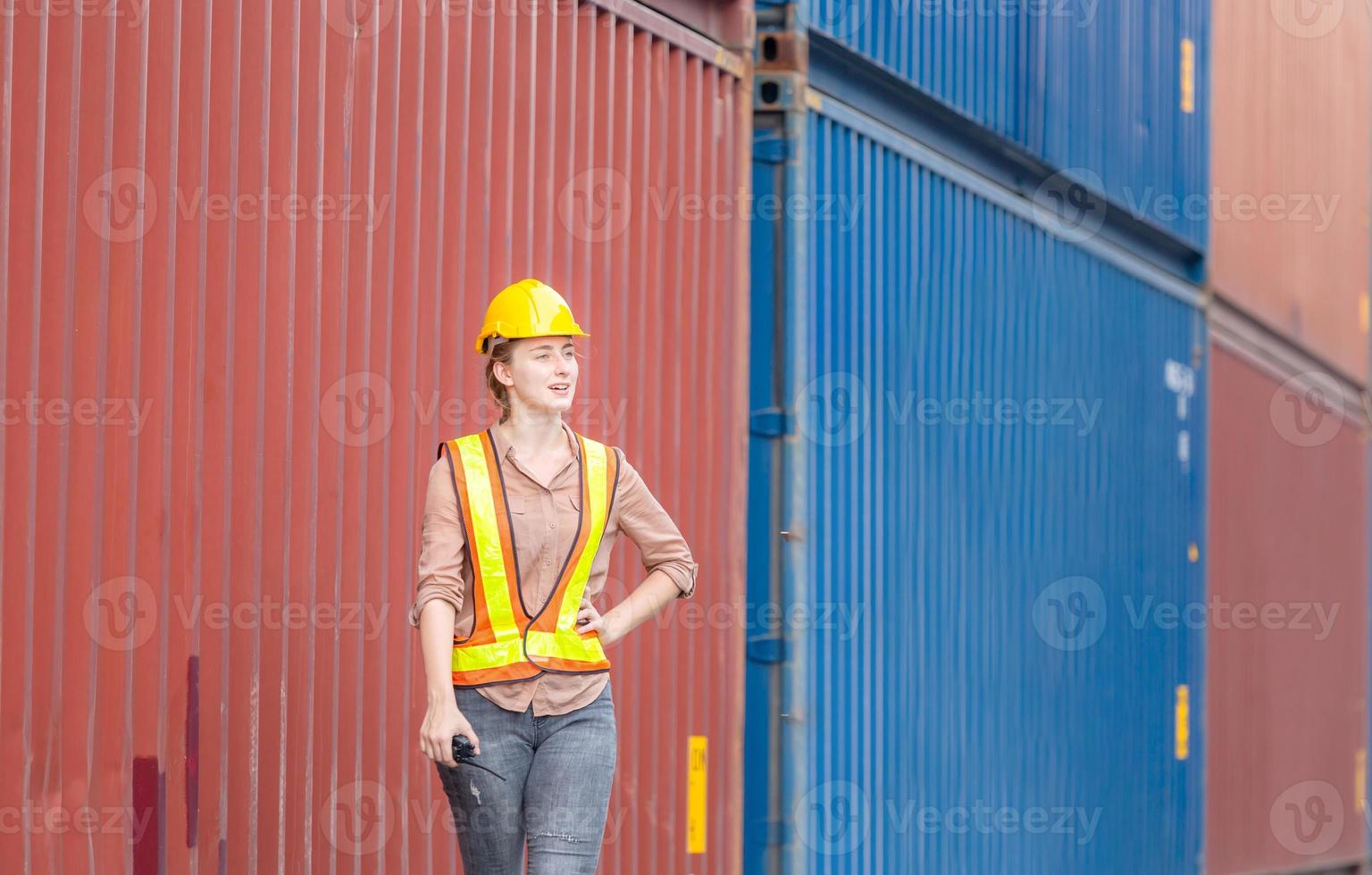 Woman engineer in hardhat and safety vest holding holding two-way radio control loading containers box from cargo photo