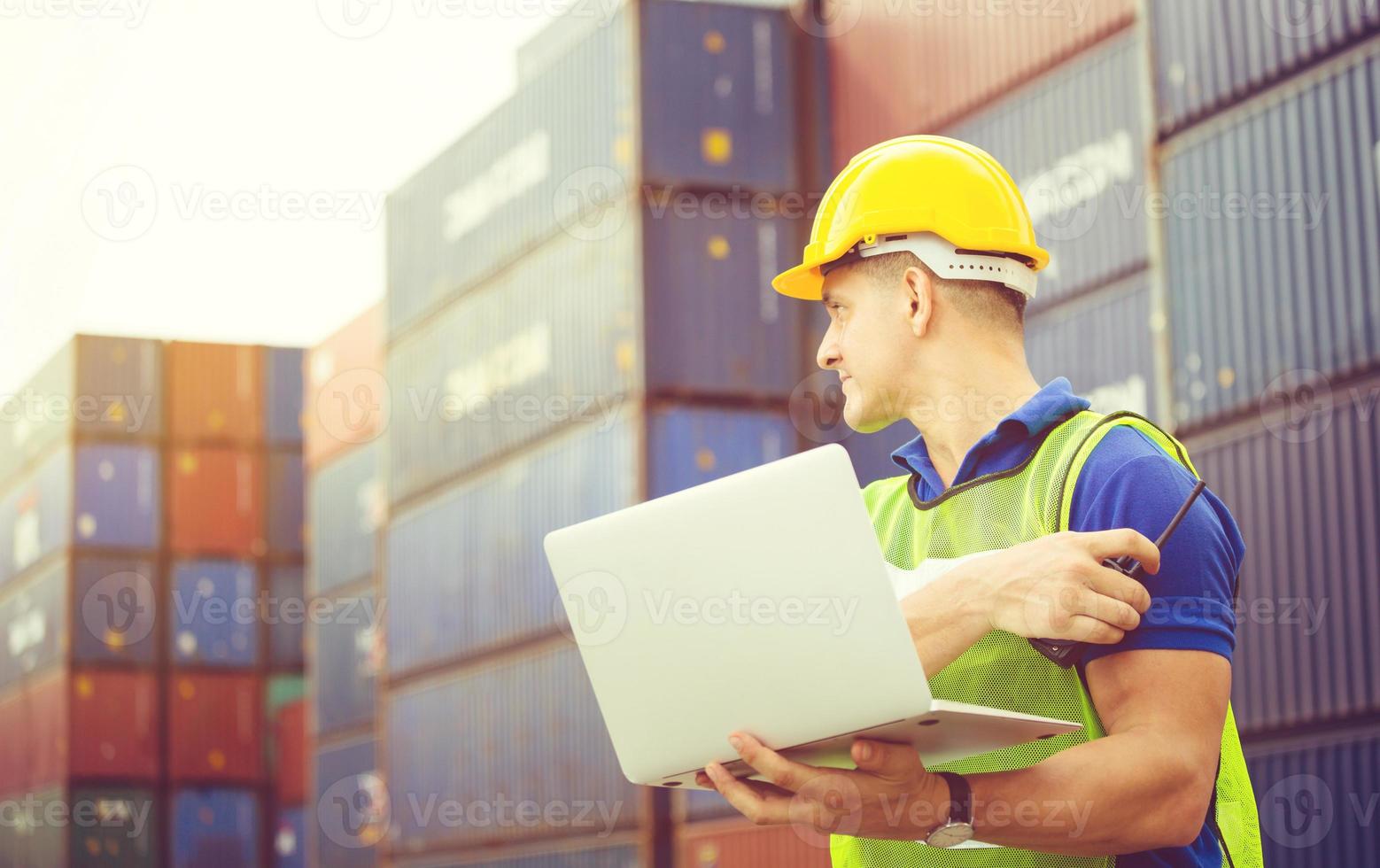 Worker man in hardhat and safety vest holding laptop and two-way radio, Foreman control loading containers box from cargo photo