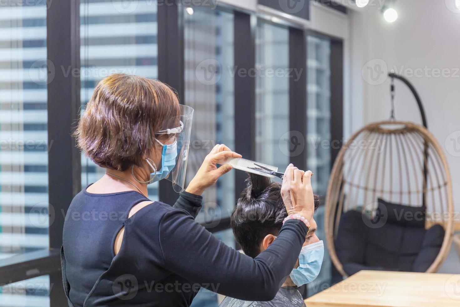 peluquera cortando el cabello del cliente con tijeras y peine, peluquera y cliente usando máscara protectora debido a la pandemia del coronavirus, nuevos conceptos normales foto