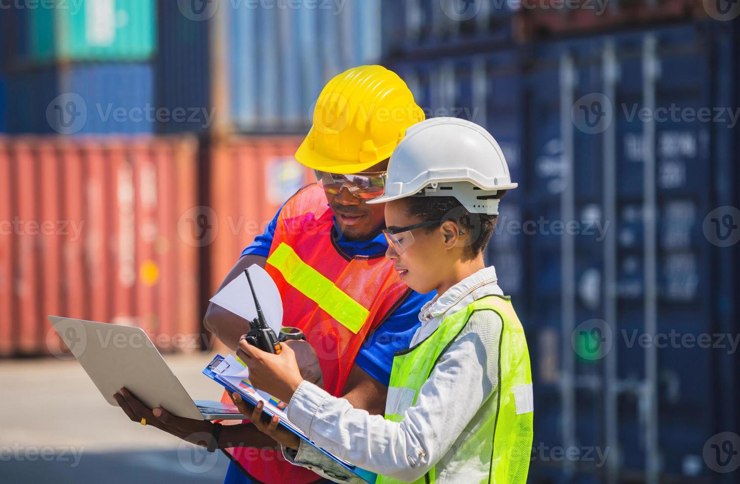 Worker man in hardhat and safety vest holding laptop and Female foreman talks on two-way radio control loading containers box from cargo photo