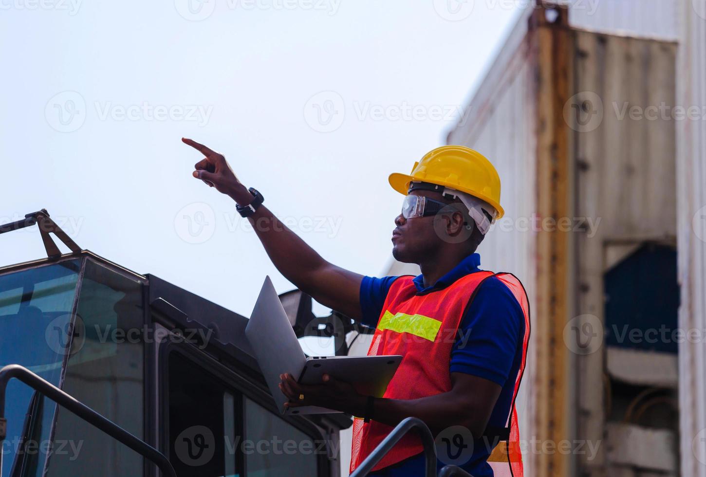 Worker man in hardhat and safety vest holding laptop standing on container stackers control loading containers box from cargo photo