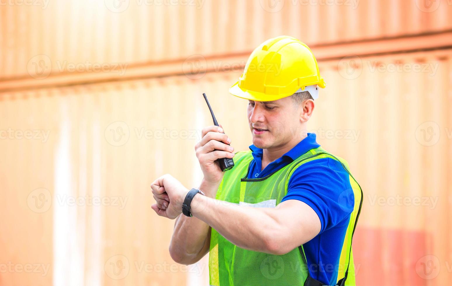 hombre ingeniero con casco y chaleco de seguridad habla por radio bidireccional, hombre trabajador mirando la hora del reloj en los contenedores de carga foto