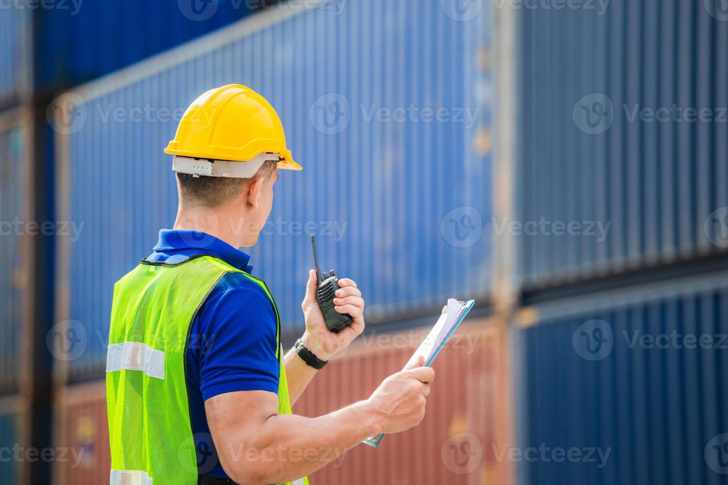 Rear view of Foreman worker in hard hat and safety vest holding clipboard checklist and talks on two-way radio at containers cargo photo