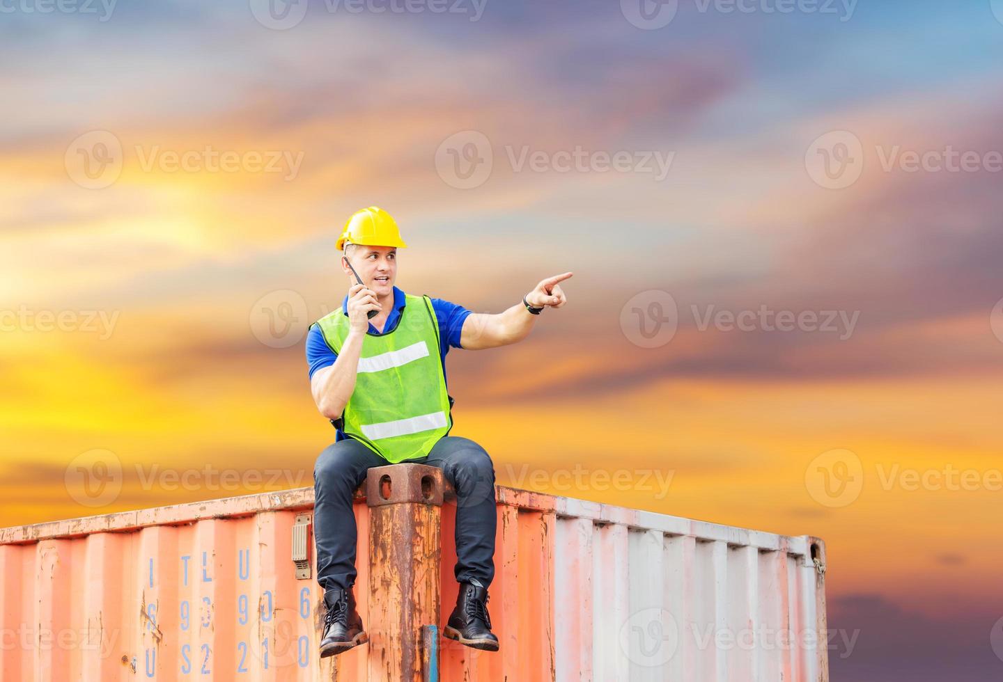 Foreman in hardhat and safety vest sitting on container box with clipping path talks on two-way radio control loading containers box from cargo photo
