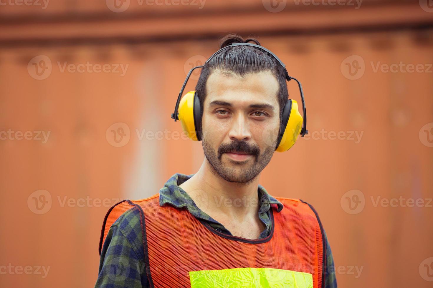 Cheerful factory worker man wearing noise reduction ear muffs and looking at camera with joy containers box background, Happiness and protection concept photo