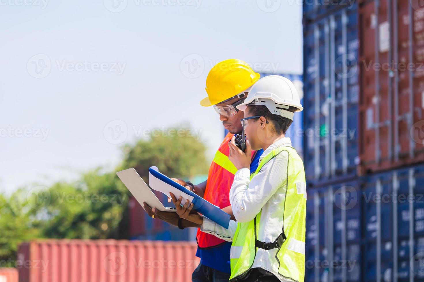 Worker man in hardhat and safety vest holding laptop and Female foreman talks on two-way radio control loading containers box from cargo photo