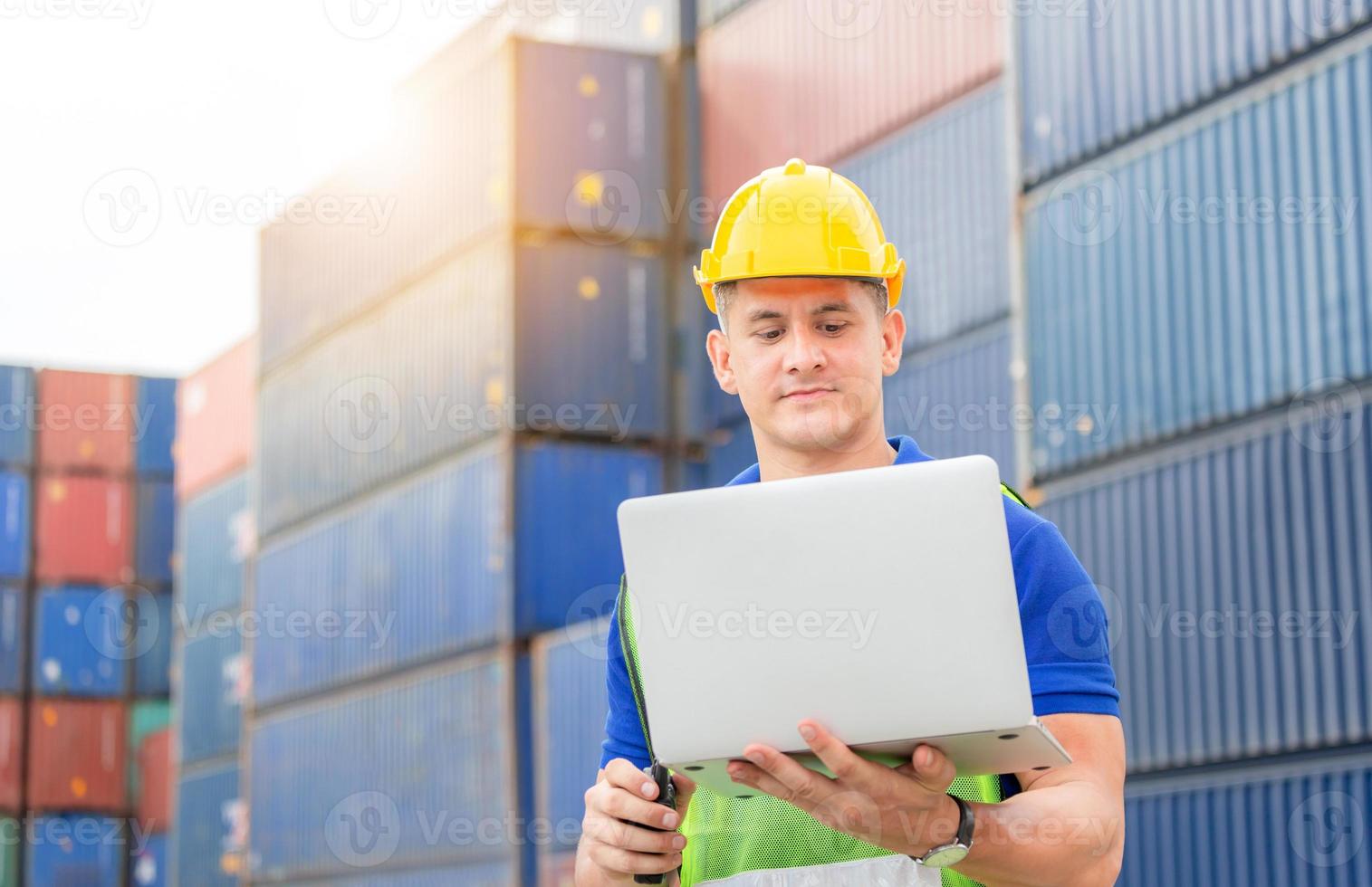 Engineer man in hardhat and safety vest holding laptop and two-way radio, Foreman worker control loading containers box from cargo photo