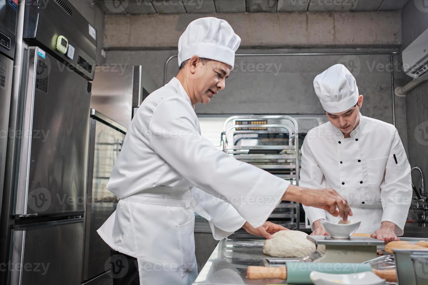 Two professional Asian male chefs in white cook uniforms and aprons are kneading pastry dough and eggs, preparing bread and fresh bakery food, baking in oven at stainless steel kitchen of restaurant. photo