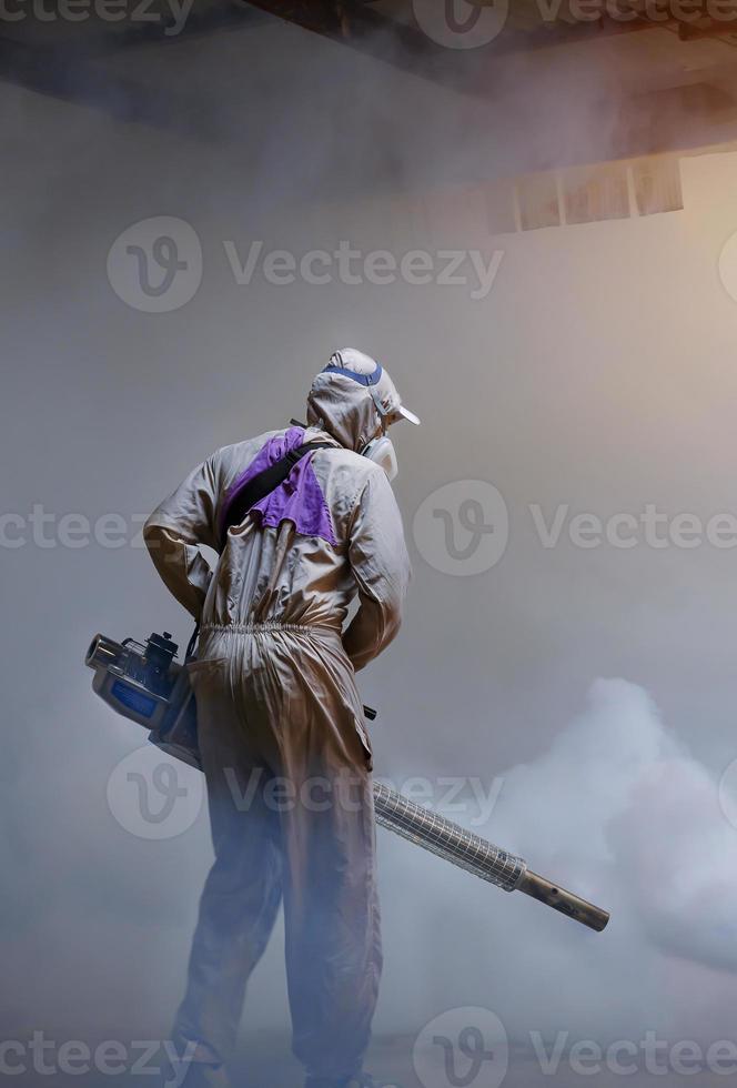 Rear view of healthcare worker using fogging machine spraying chemical to eliminate mosquitoes inside of storage at slum area photo