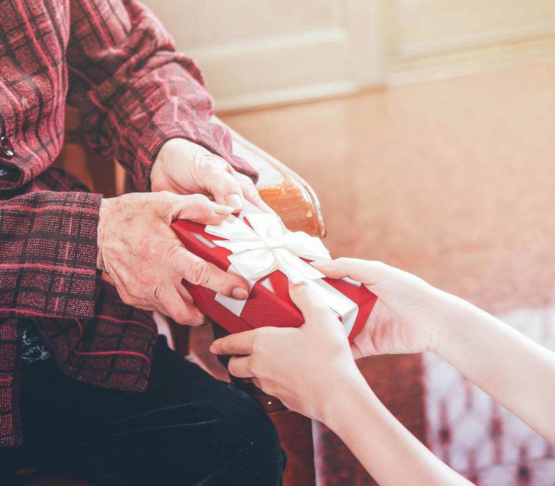 Teenage asia girl is giving grandma sitting on chair beautiful giftbox for mothers day birthday celebration idea at home with dark background, close up, copy space, cropped view photo