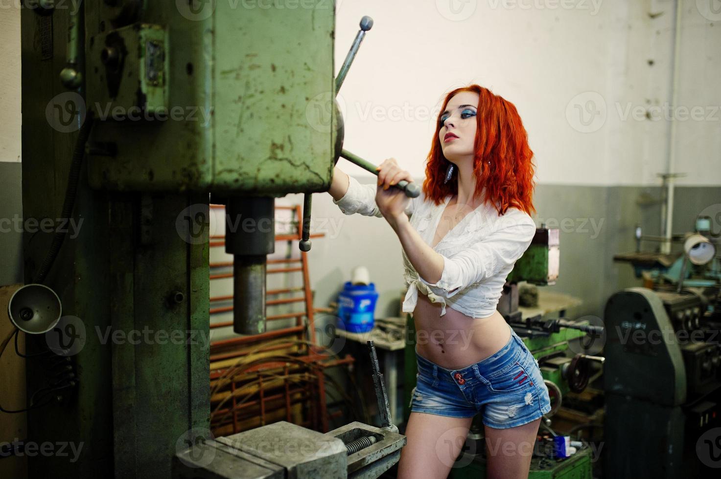 Red haired girl wear on short denim shorts and white blouse posed at industrial machine at the factory. photo