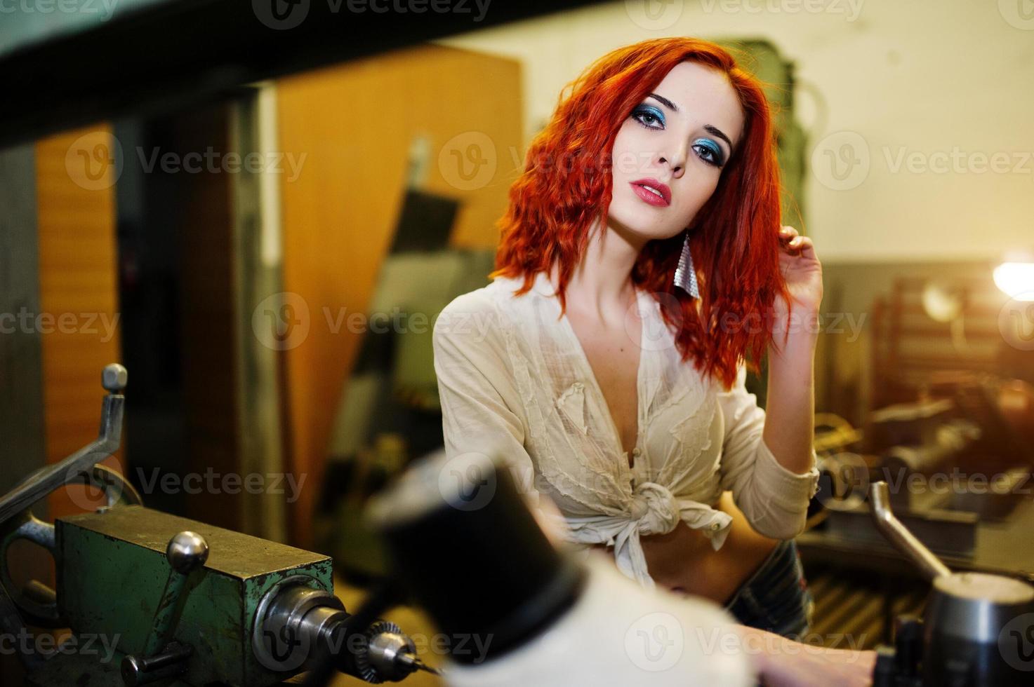 Red haired girl wear on short denim shorts and white blouse posed at industrial machine at the factory. photo