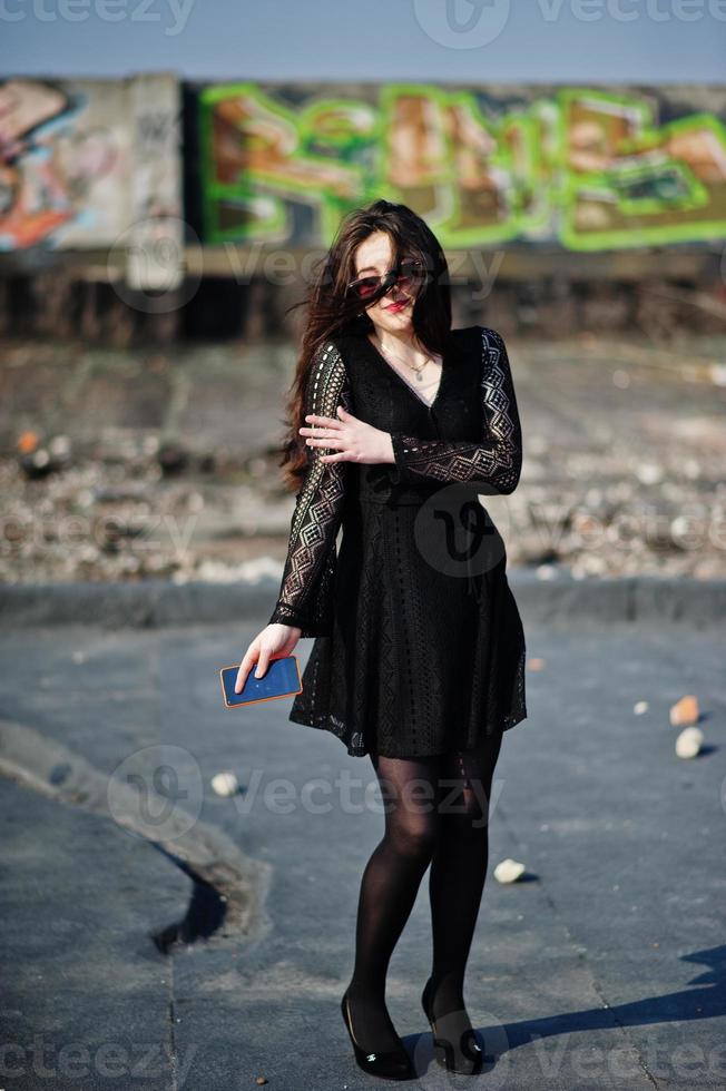 retrato de una chica morena con labios rojos y teléfono móvil naranja en las manos, con un vestido negro, gafas de sol posadas en el techo. modelo de moda callejera. foto