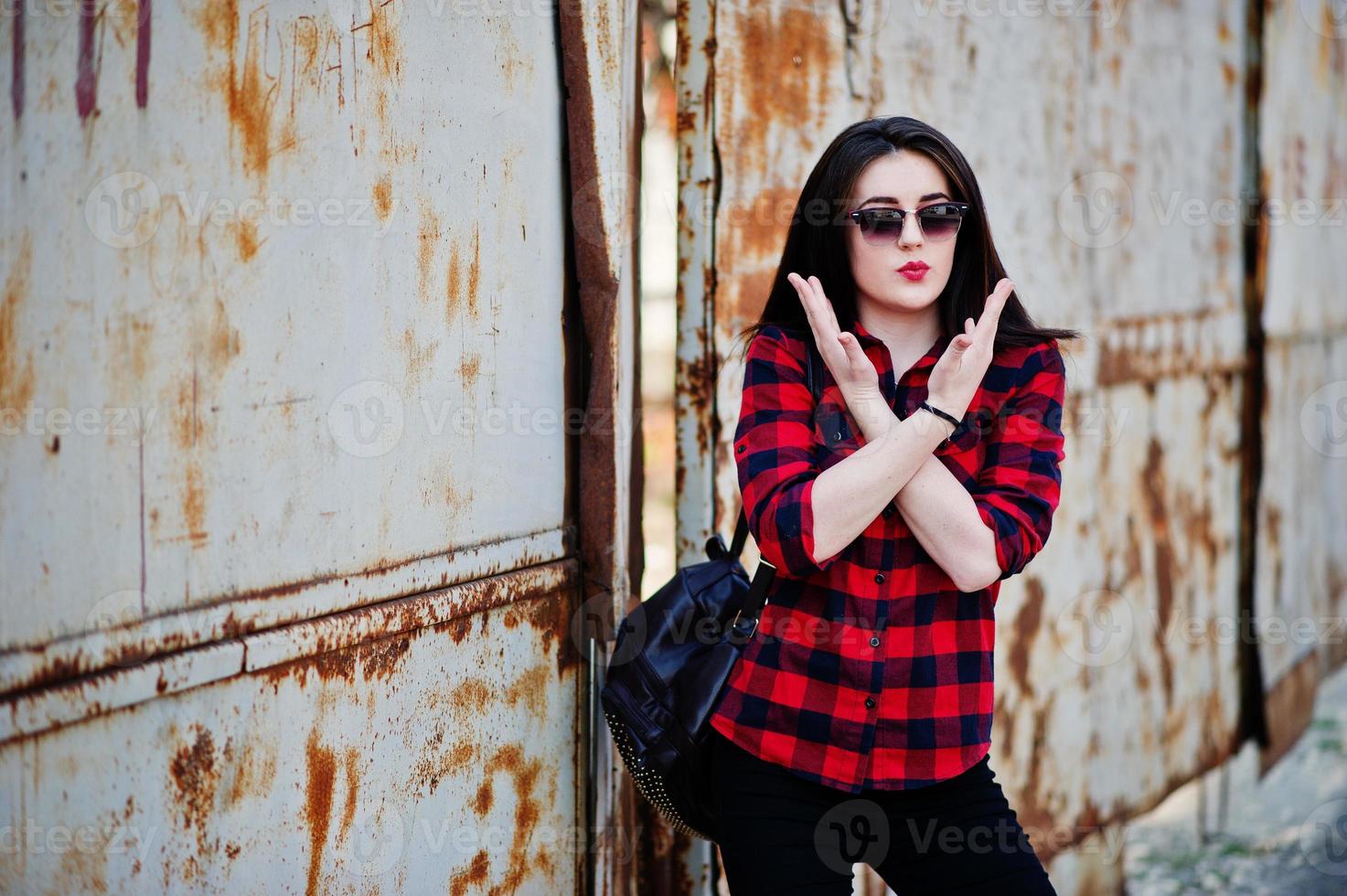 Fashion portrait girl with red lips wearing a red checkered shirt, sunglasses and backpack background rusty fence. photo