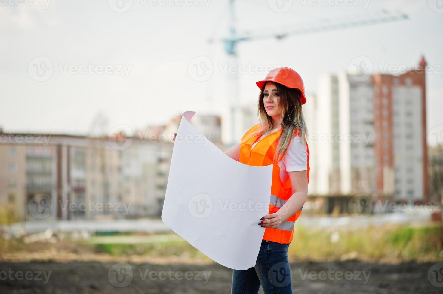 Engineer builder woman in uniform waistcoat and orange protective helmet hold business paper against new buildings with crane. photo