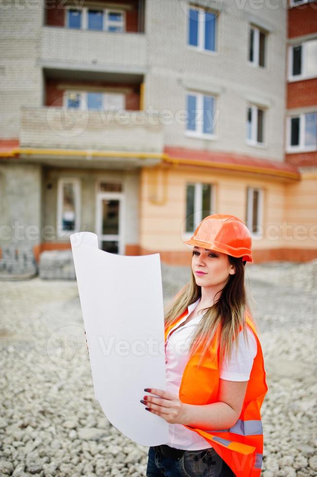 mujer ingeniera constructora con chaleco uniforme y casco protector naranja sostiene papel comercial contra el nuevo edificio. tema de bloque de vivienda de propiedad. foto