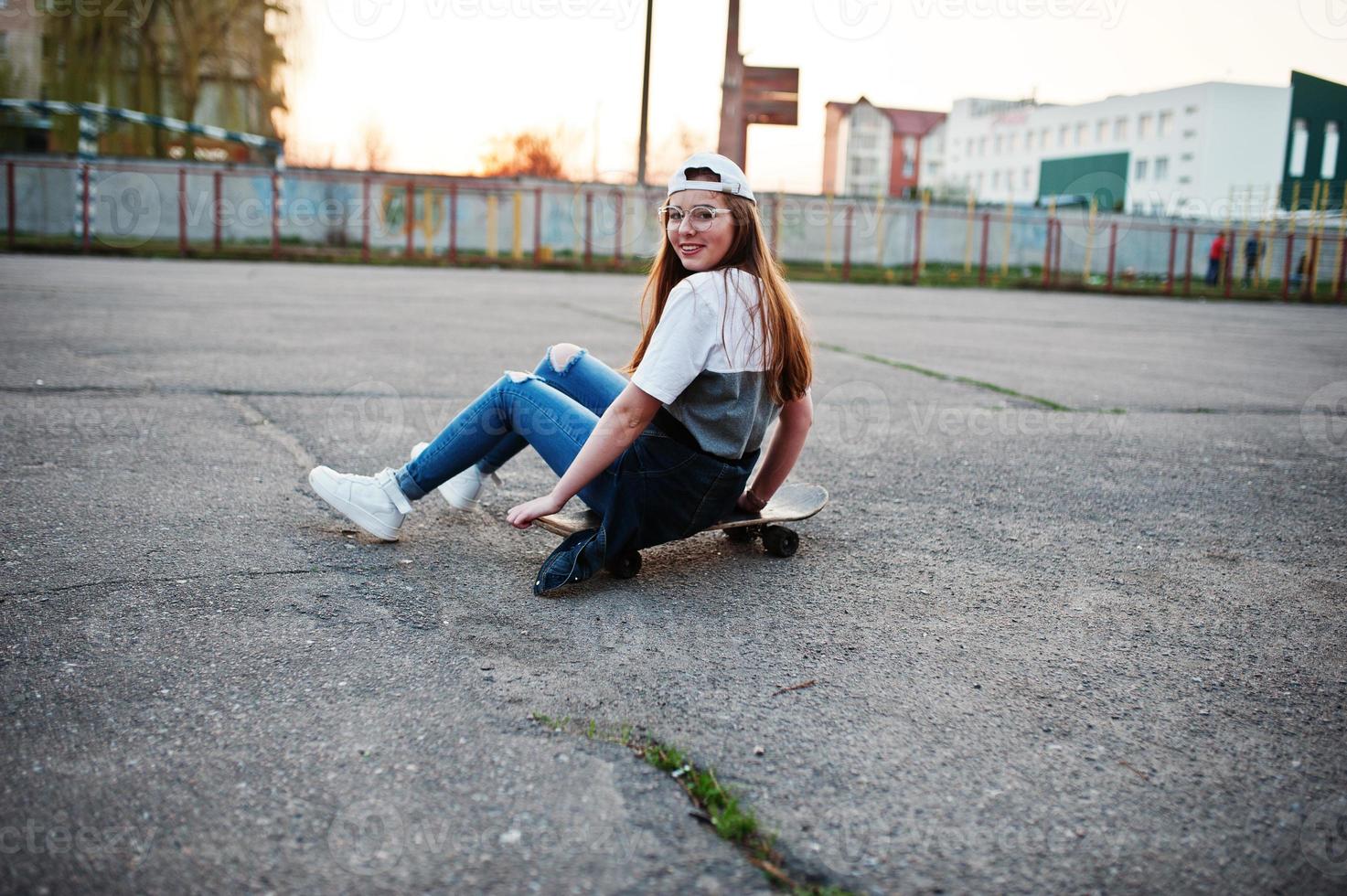 joven adolescente urbana con monopatín, gafas, gorra y jeans rotos en el campo de deportes del patio al atardecer. foto