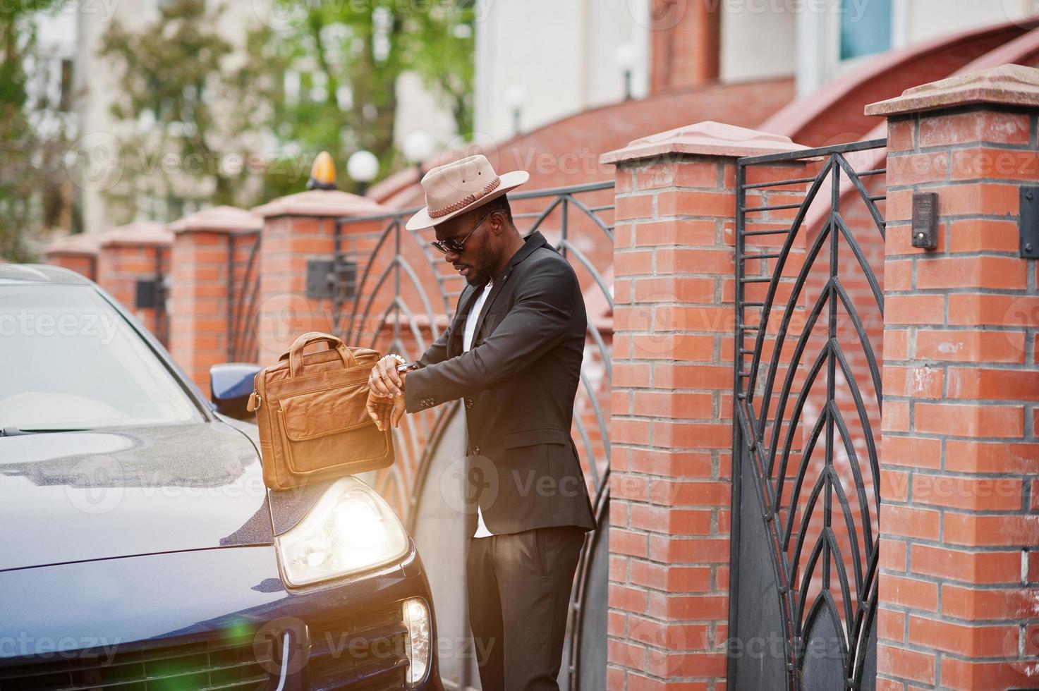 Stylish black man at glasses with hat, wear on suit with handbag against luxury car. Rich african american businessman. Look at watches. photo