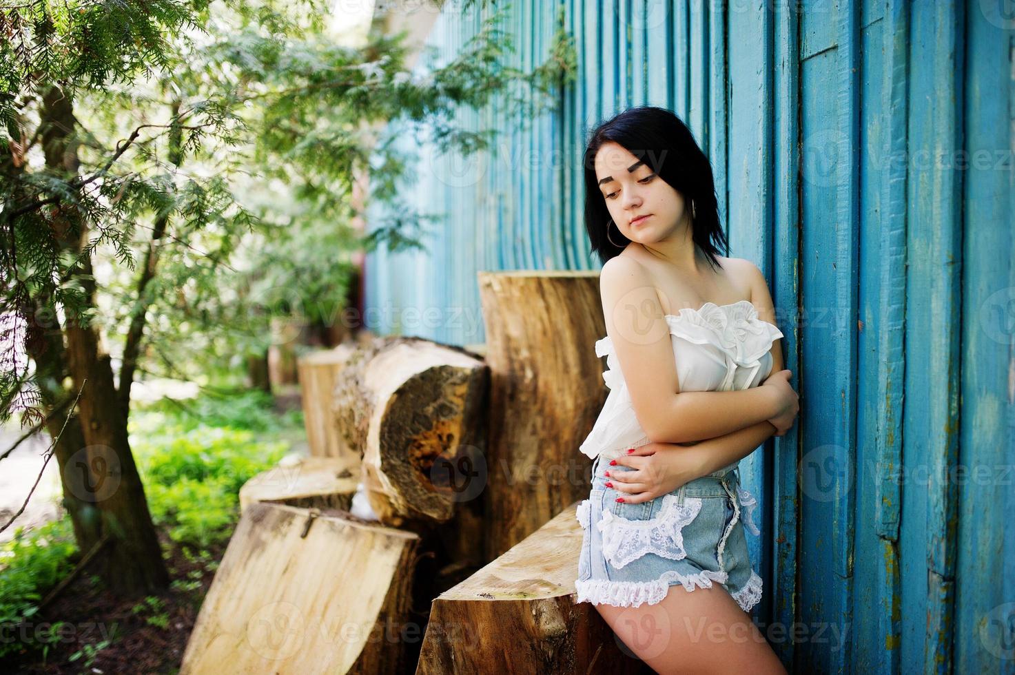 retrato de chica morena sexy en shorts de jeans de mujer y blusa blanca contra casa de madera azul con tocones. foto
