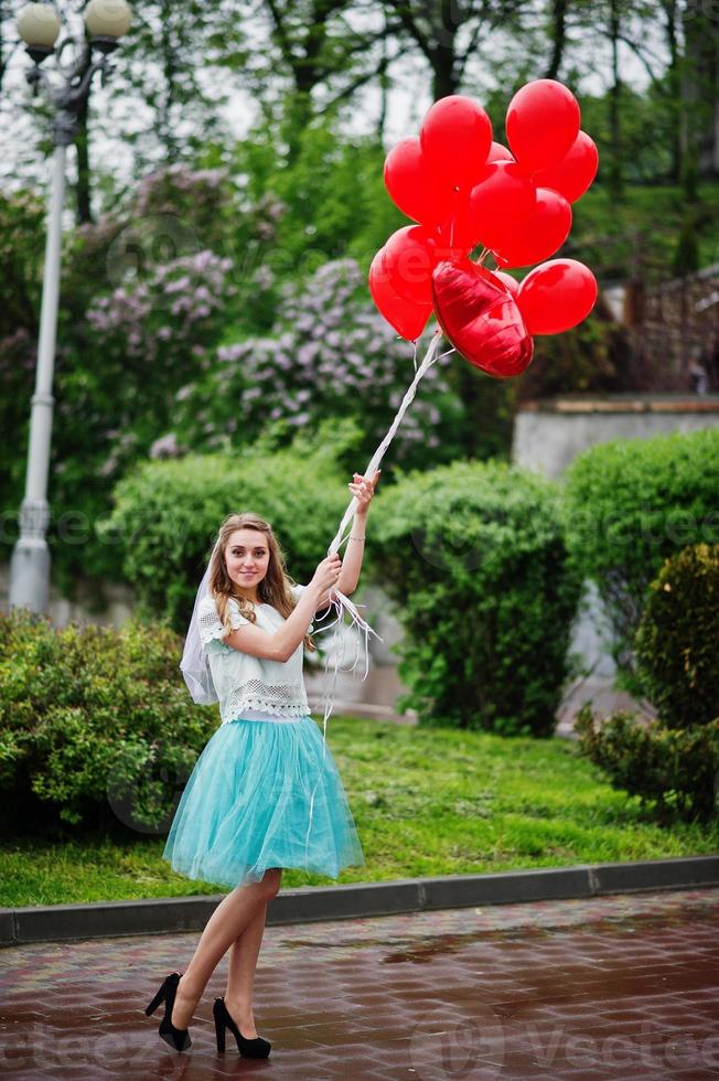 Portrait of a gorgeous beautiful bride wearing pretty dress and veil holding heart-shaped red balloons in the park at bachelorette party. photo