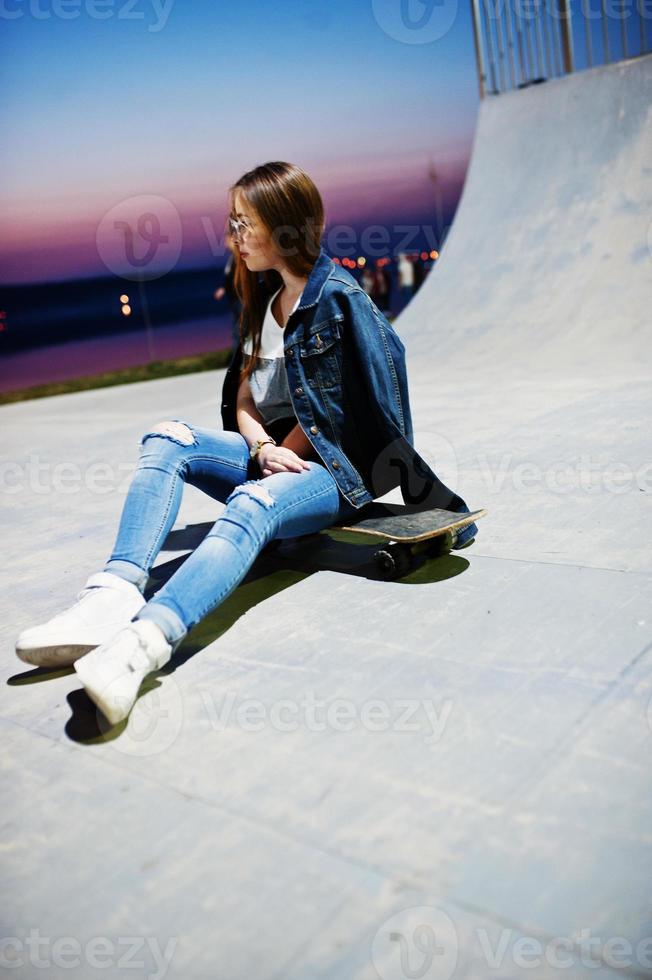Young teenage urban girl with skateboard, wear on glasses, cap and ripped jeans at skate park on the evening. photo