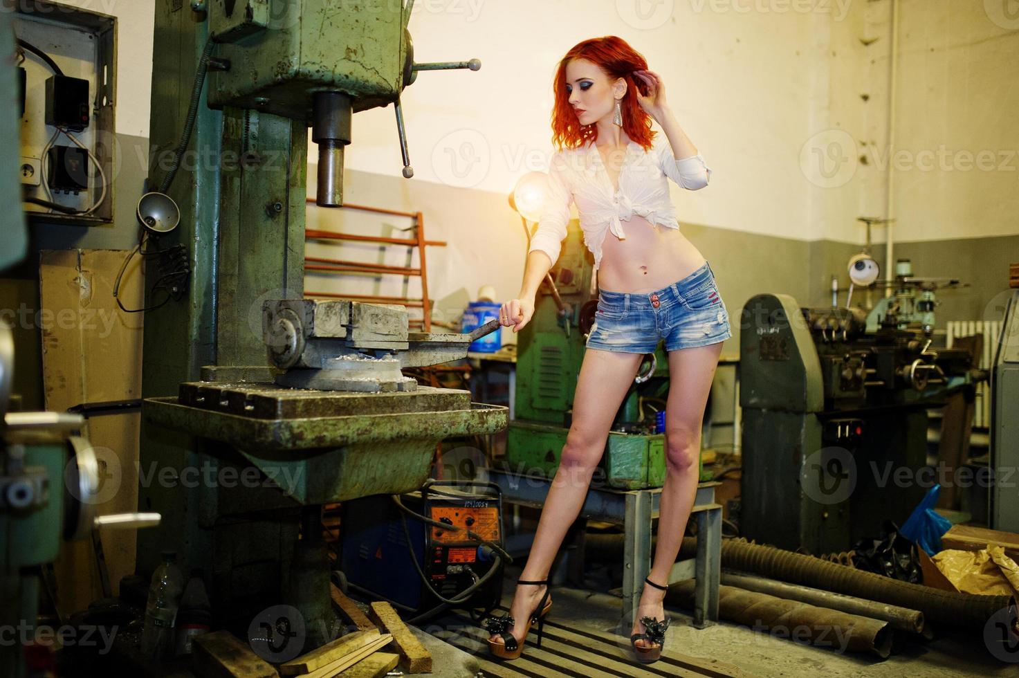 Red haired girl wear on short denim shorts and white blouse posed at industrial machine at the factory. photo