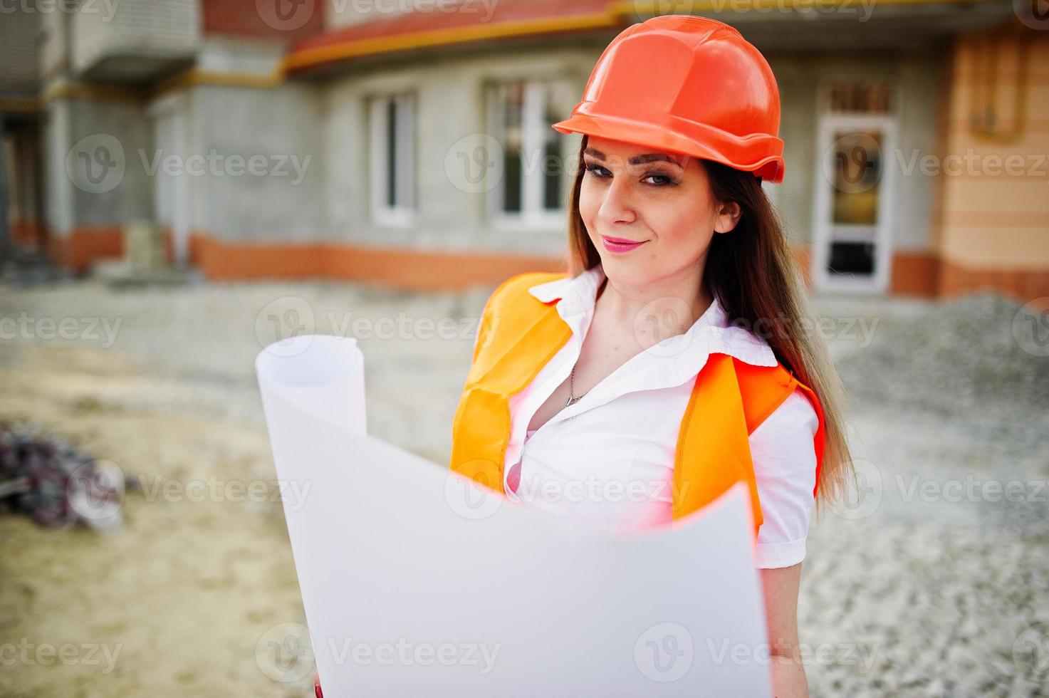 Engineer builder woman in uniform waistcoat and orange protective helmet hold business paper against new building. Property living block theme. photo