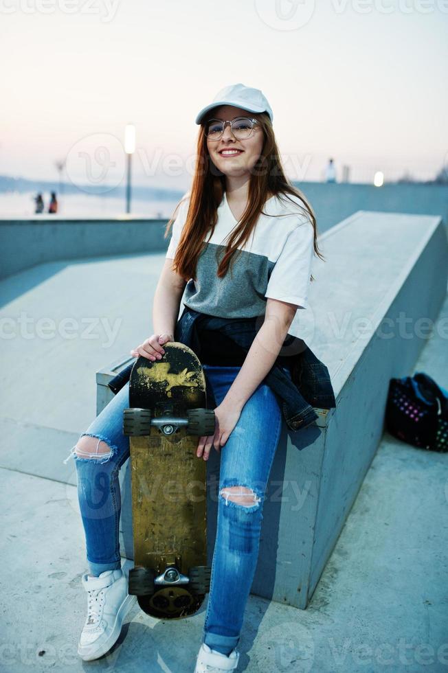 joven adolescente urbana con monopatín, gafas, gorra y jeans rotos en el parque de patinaje por la noche. foto