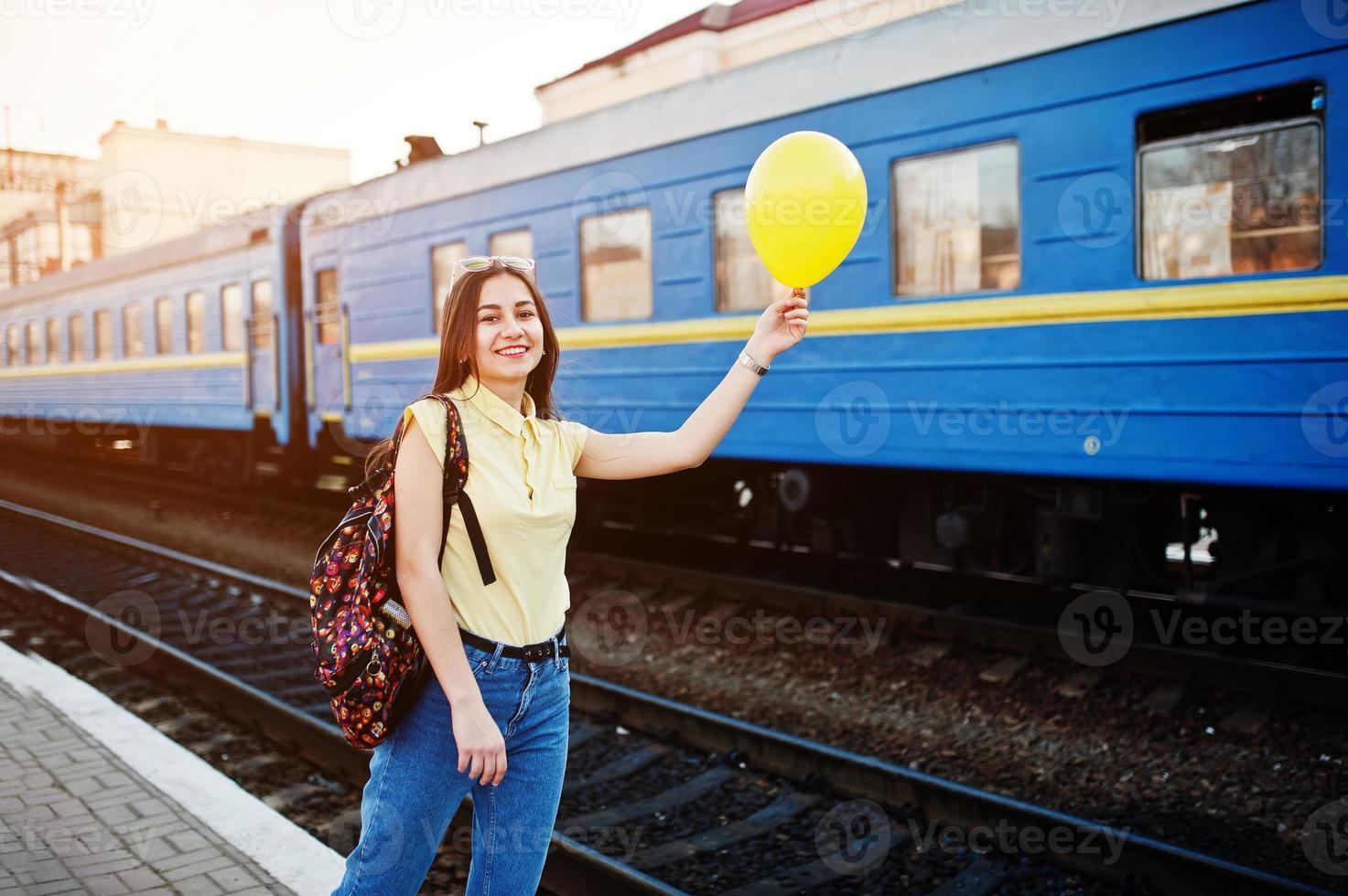 joven adolescente de pie en el andén de la estación de tren con globo a mano, vestida con camiseta amarilla, jeans y gafas de sol, con mochila. foto