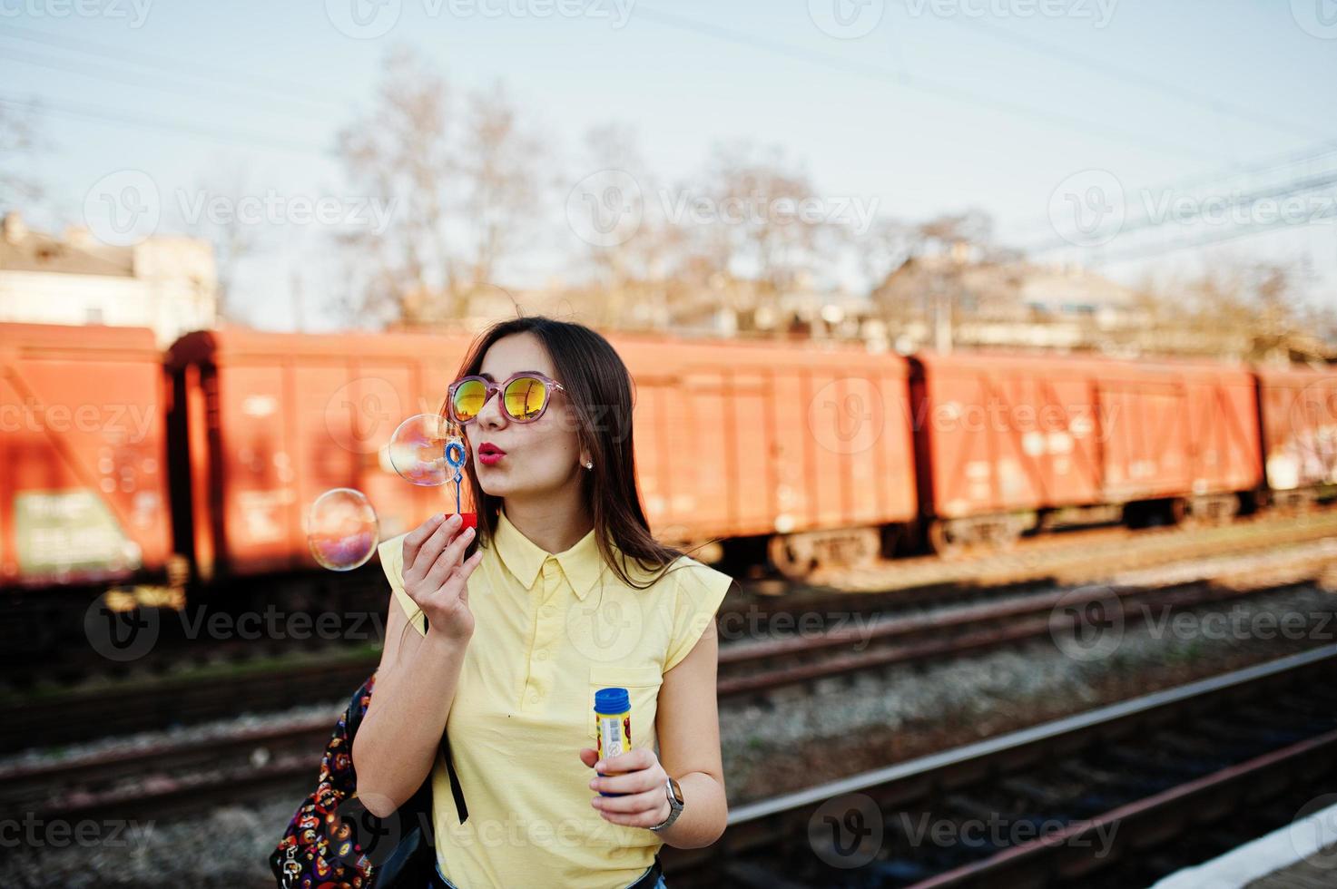 joven adolescente de pie en el andén de la estación de tren y soplando burbujas de jabón, vestida con camiseta amarilla, jeans y gafas de sol, con mochila. foto