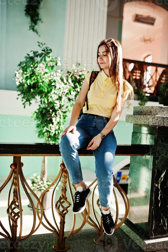 joven adolescente sentada en las barandillas de las escaleras, vestida con una camiseta amarilla, jeans y gafas de sol con mochila. foto