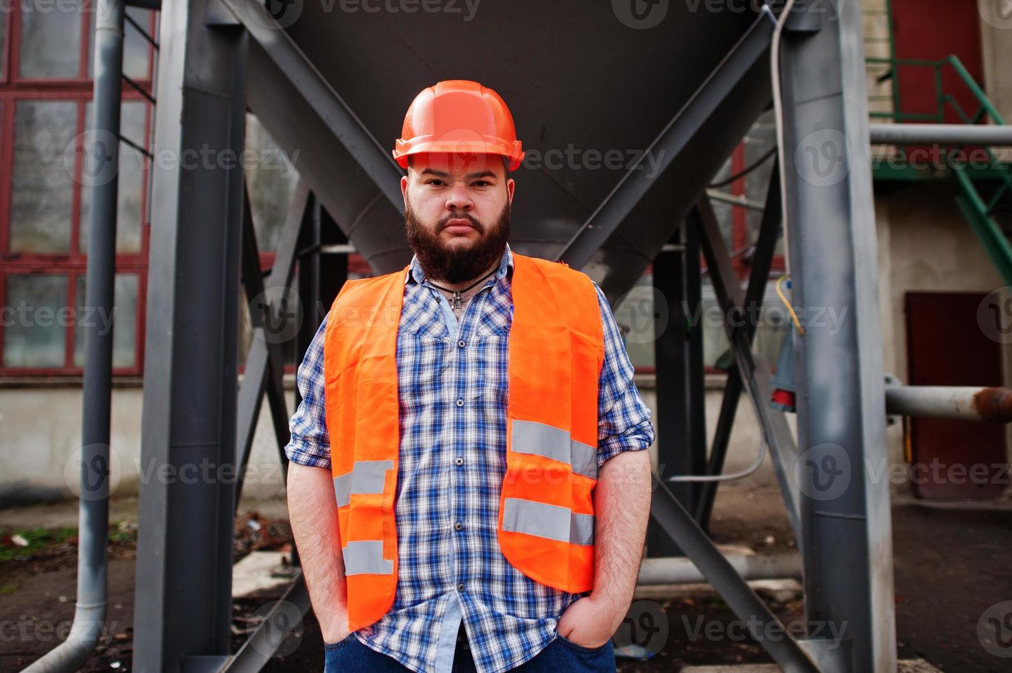 trabajador de barba brutal traje de hombre trabajador de la construcción en casco naranja de seguridad quédese cerca de un gran barril industrial. foto