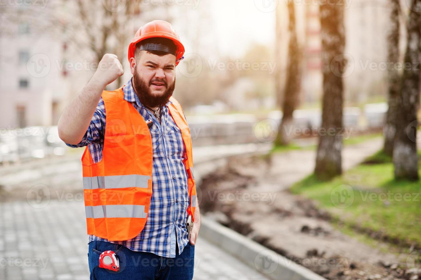 Portrait of brutal angry beard worker man suit construction worker in safety orange helmet against pavement with showing arms. photo